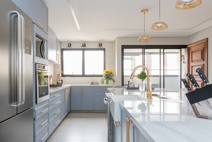 Projeto Mari Milani| Cozinha ampla com ilha central e marcenaria em azul claro. A área de serviço fica integrada, mas discreta, graças à porta de serralheria com vidro canelado, garantindo luz natural e organização.